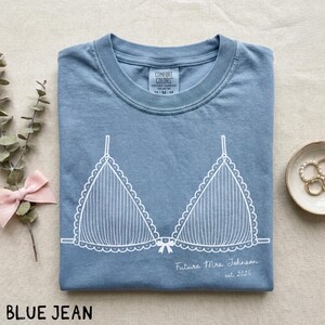 Puede incluir: Camiseta azul vaquero con un gr&aacute;fico blanco de sujetador. El texto "Future Mrs. Johnson est. 2026" est&aacute; impreso debajo del dise&ntilde;o del sujetador. La camiseta est&aacute; doblada cuidadosamente sobre una superficie neutra.