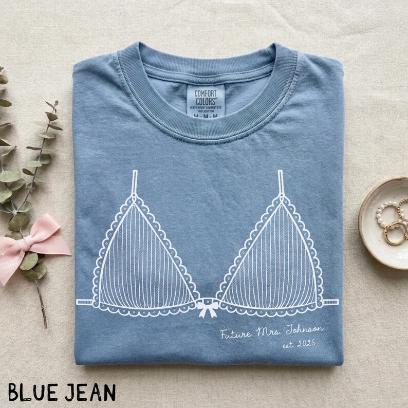 Puede incluir: Camiseta azul vaquero con un gr&aacute;fico blanco de sujetador. El texto "Future Mrs. Johnson est. 2026" est&aacute; impreso debajo del dise&ntilde;o del sujetador. La camiseta est&aacute; doblada cuidadosamente sobre una superficie neutra.