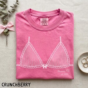Puede incluir: Una camiseta rosa Comfort Colors con un gr&aacute;fico blanco de sujetador. La camiseta de cuello redondo tiene mangas cortas. El texto "Miami, FL est. 2026" est&aacute; impreso en la camiseta. La palabra "CRUNCHBERRY" est&aacute; impresa en la parte inferior.