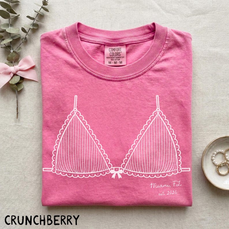 Puede incluir: Una camiseta rosa Comfort Colors con un gr&aacute;fico blanco de sujetador. La camiseta de cuello redondo tiene mangas cortas. El texto "Miami, FL est. 2026" est&aacute; impreso en la camiseta. La palabra "CRUNCHBERRY" est&aacute; impresa en la parte inferior.