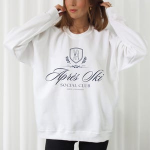 May include: White sweatshirt with a navy blue logo that says "Après Ski Social Club Aspen, Colorado" with a graphic of crossed skis inside a shield.