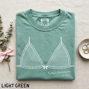 Puede incluir: Camiseta verde claro con un gr&aacute;fico blanco de un sujetador. La camiseta tiene el texto "Kailey's Bachelorette est. 2026" impreso. La camiseta est&aacute; doblada cuidadosamente. La etiqueta de la marca "Comfort Colors" es visible.