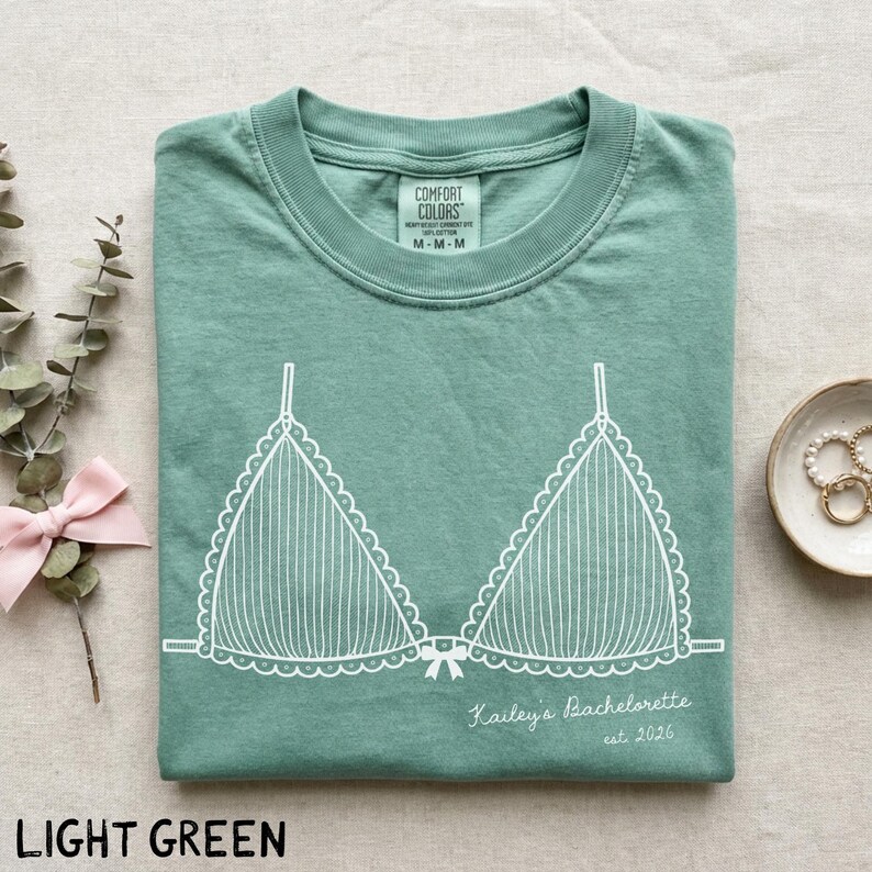 Puede incluir: Camiseta verde claro con un gr&aacute;fico blanco de un sujetador. La camiseta tiene el texto "Kailey's Bachelorette est. 2026" impreso. La camiseta est&aacute; doblada cuidadosamente. La etiqueta de la marca "Comfort Colors" es visible.