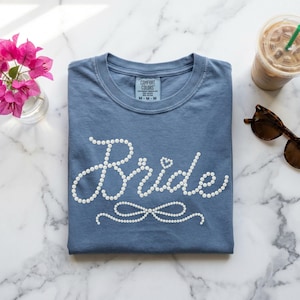 May include: A blue t-shirt with the word "Bride" in white, embellished with small, round details. The shirt is folded neatly on a marble surface, with a small vase of pink flowers, sunglasses, and a drink nearby.
