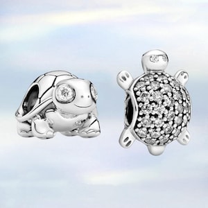Può includere: Due ciondoli a forma di tartaruga in argento, uno con un guscio dettagliato e l'altro con un guscio liscio e pietre chiare scintillanti.