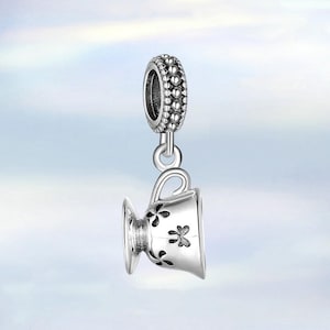 May include: A silver charm in the shape of a teacup, with a decorative floral design. The charm has a loop at the top for attaching to a bracelet or necklace. The charm is suspended from a beaded ring.