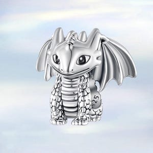 May include: A silver-colored dragon figurine with large wings, a smiling face, and detailed scales. The dragon is sitting upright, with its wings spread slightly. The figurine has a polished finish and is set against a soft, blurred background.