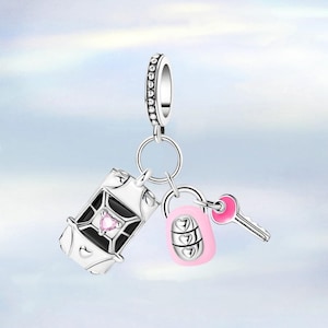 Könnte beinhalten: Ein silberner Charm-Anhänger mit einer Reisetasche, einem Zahlenschloss und einem Schlüssel. Die Truhe hat ein schwarz-weißes Design mit einem rosa herzförmigen Edelstein. Schloss und Schlüssel sind rosa.