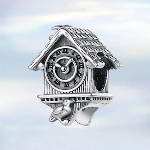 Könnte beinhalten: Ein silberfarbener Charm in Form einer Kuckucksuhr. Die Uhr hat ein rundes Zifferblatt mit römischen Ziffern, einen Vogel darunter und ein strukturiertes Dach. Der Charm ist mit vertikalen Linien und einem dekorativen Rand versehen.