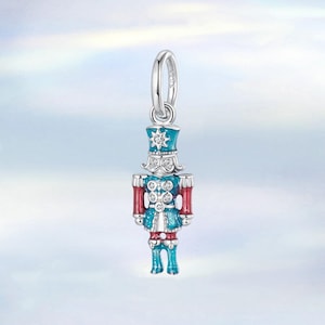 May include: A silver charm depicting a toy soldier nutcracker. The charm is painted in blue, red, and white, with a star design on the hat. It has a loop at the top for attaching to a bracelet or necklace. A festive accessory.