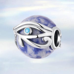 May include: A silver-colored charm with an eye design, featuring a blue gemstone in the center and a blue and white marbled sphere. The charm has an open design and is likely for a bracelet or necklace.