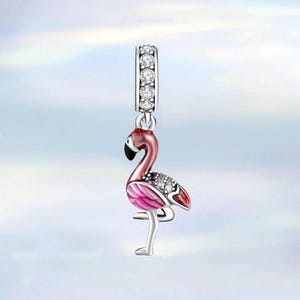 Könnte beinhalten: Ein Flamingo-Charm mit einer silberfarbenen Schlaufe, die mit klaren Kristallen besetzt ist. Der rosa Flamingo hat einen schwarzen Schnabel und ein silberfarbenes Bein und steht auf einem Bein.