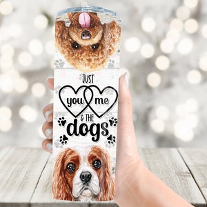 May include: A stainless steel tumbler featuring watercolor illustrations of two dogs and paw prints. The tumbler has the text "Just you & me & the dogs" in a black font, with hearts and paw prints.