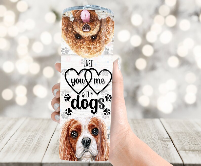 May include: A stainless steel tumbler featuring watercolor illustrations of two dogs and paw prints. The tumbler has the text "Just you & me & the dogs" in a black font, with hearts and paw prints.