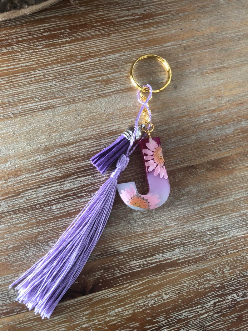 Peut inclure: Porte-cl&eacute;s en r&eacute;sine lettre J violet et blanc avec des fleurs press&eacute;es et un pompon violet.
