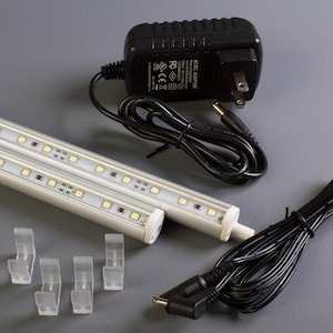 May include: Two white LED light strips with a black power adapter, a black power cord, and four clear plastic mounting clips.