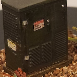 May include: A miniature model of a green electrical box with a warning sign and a small sign that says "EVLSG". The box is sitting on a bed of brown and white gravel.