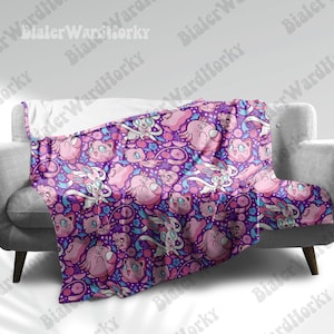 May include: A purple blanket featuring a repeating pattern of pink cartoon characters, flowers, and swirls. The blanket is draped over a gray sofa. The characters include round, cat-like creatures and others with long ears and wings.