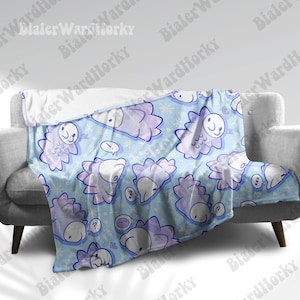 May include: A light blue blanket with a repeating pattern of cartoon-like creatures in shades of purple and white. The blanket has a soft, plush texture and is draped over a gray sofa. The creatures have speech bubbles and snowflake accents.