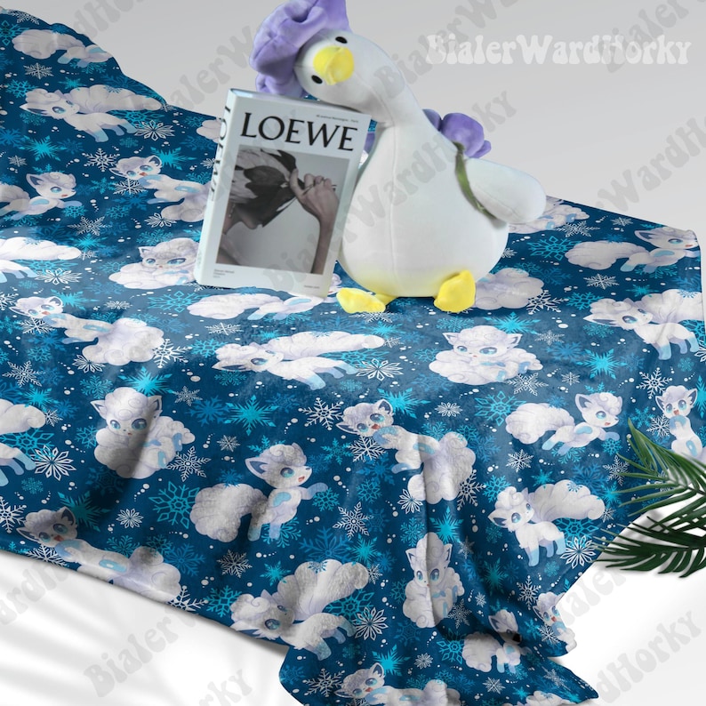 May include: A blue blanket featuring a pattern of white cartoon foxes and snowflakes. A plush goose toy with a purple head and yellow feet rests on the blanket, alongside a book with "LOEWE" on the cover.