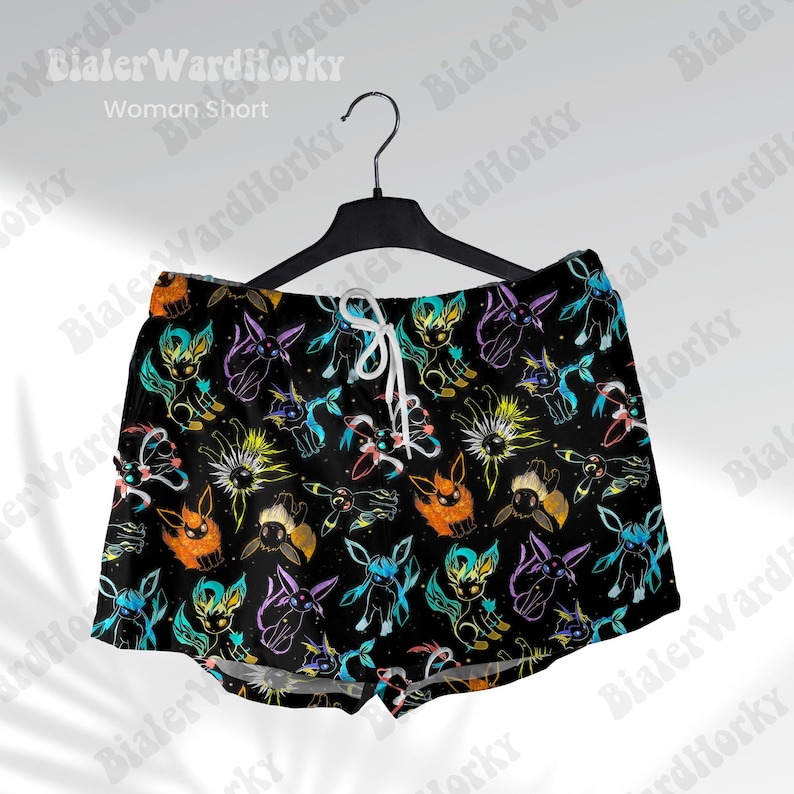 Puede incluir: Shorts negros con un patr&oacute;n colorido y repetitivo de criaturas de dibujos animados. Los shorts tienen una cintura con cord&oacute;n y cuelgan de una percha negra. El texto "Woman Short" es visible en la parte superior.