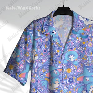 May include: A short-sleeved shirt with a vibrant, repeating pattern. The shirt's design includes cartoon characters, musical notes, hearts, and flowers in pink, yellow, and blue on a light blue background. The shirt is displayed on a black hanger.