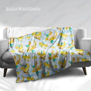 May include: A light blue blanket featuring a repeating pattern of yellow cartoon dragons with green wings. The blanket is draped over a grey sofa, with a white fluffy underside visible.
