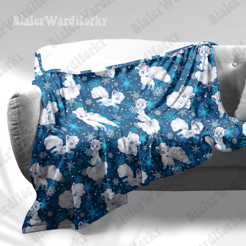 May include: A blue blanket with a repeating pattern of white cartoon foxes and snowflakes. The blanket is draped over a grey sofa. The foxes have large eyes and fluffy tails, and the snowflakes are in shades of blue and white.