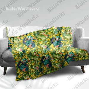 Peut inclure: Une couverture à motifs verts, bleus et jaunes, drapée sur un canapé gris. Le motif présente des créatures stylisées répétées et des éléments floraux jaunes sur un fond vert foncé. La couverture semble être faite d'une matière douce.