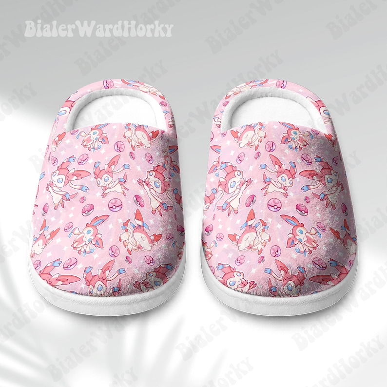 Może przedstawiać: Para r&oacute;żowych i białych pluszowych kapci. Kapcie mają powtarzający się wz&oacute;r postaci z kresk&oacute;wek, r&oacute;żowych serc i pokeballi. Kapcie mają białą podeszwę i białe wnętrze.