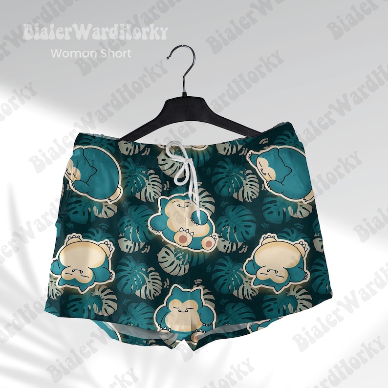 May include: Teal and beige shorts with a pattern of cartoon characters and tropical leaves. The shorts are hanging on a black hanger. The text "Woman Short" is visible at the top.