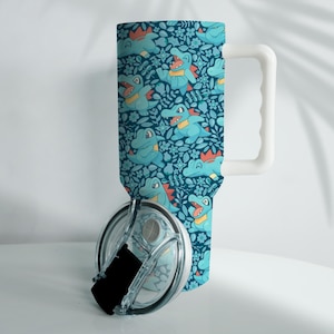 May include: A tumbler with a white handle and a clear lid. The tumbler features a repeating pattern of cartoon alligators in various poses, with orange and yellow accents, set against a dark teal floral background.