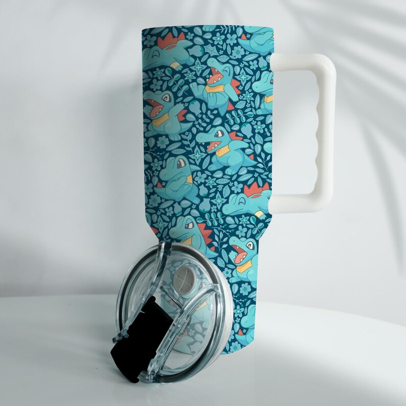 May include: A tumbler with a white handle and a clear lid. The tumbler features a repeating pattern of cartoon alligators in various poses, with orange and yellow accents, set against a dark teal floral background.