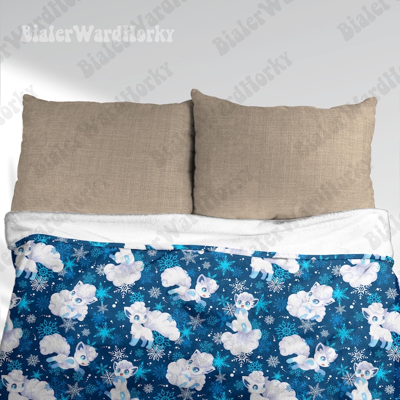 May include: A blue blanket with a white sherpa lining, patterned with white cartoon foxes and snowflakes. Two beige pillows are positioned above the blanket, creating a cosy bedding arrangement.