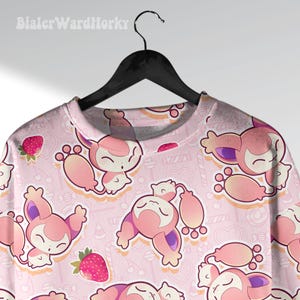 May include: Pink crew neck shirt with an all-over pattern of cartoon creatures, strawberries, and candies. The creatures are pink and white with purple accents. The shirt is hanging on a black hanger.