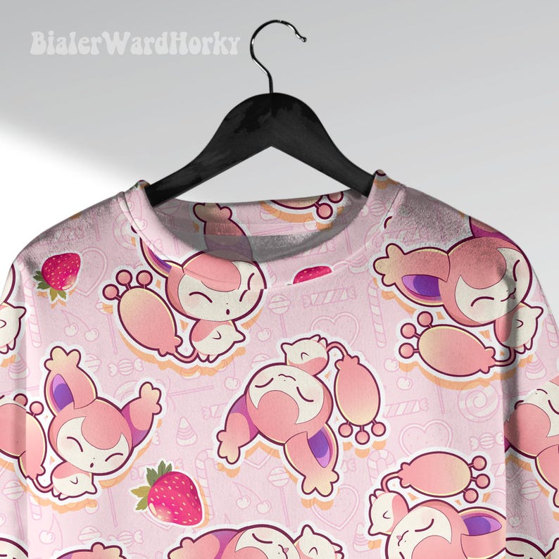 May include: Pink crew neck shirt with an all-over pattern of cartoon creatures, strawberries, and candies. The creatures are pink and white with purple accents. The shirt is hanging on a black hanger.
