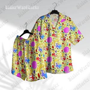 May include: A matching pajama set featuring a short-sleeved button-down shirt and shorts. The set is light yellow with a repeating pattern of cartoon turtles, purple circles, and various fruits. The shirt and shorts are hanging on a black hanger.