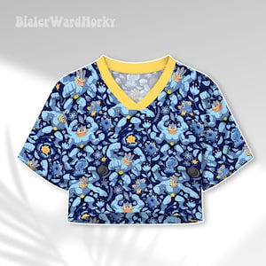 May include: A short-sleeved crop top with a V-neckline and a yellow collar. The fabric features a dark blue background with a repeating pattern of light blue four-armed creatures, yellow starbursts, and blue floral elements. The text BialerWardHorky is visible.