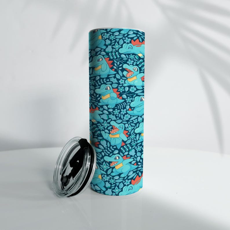 May include: A cylindrical tumbler with a teal and navy blue pattern featuring cartoon alligators and floral elements. The tumbler has a black lid and is designed for beverages.