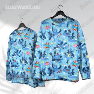 Op de afbeelding: Twee lichtblauwe sweatshirts met een herhaald patroon van stripfiguren, tekstballonnen en stripgeluidseffecten. De sweatshirts hangen aan zwarte hangers tegen een witte achtergrond.