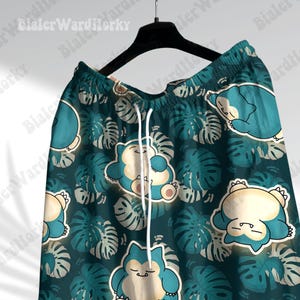 K&ouml;nnte beinhalten: Gr&uuml;n-blaue gemusterte Shorts mit einem Cartoon-Charakter-Druck. Der Charakter ist eine blaue und wei&szlig;e Kreatur mit einem gro&szlig;en Kopf und einem kleinen K&ouml;rper. Die Shorts haben einen Kordelzugbund und eine lockere Passform.