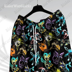 Puede incluir: Pantalones cortos negros con un patr&oacute;n colorido de varias criaturas de dibujos animados. Las criaturas est&aacute;n delineadas en colores brillantes que incluyen azul, naranja, amarillo y morado. Los pantalones cortos tienen un cord&oacute;n blanco y cuelgan de una percha negra.