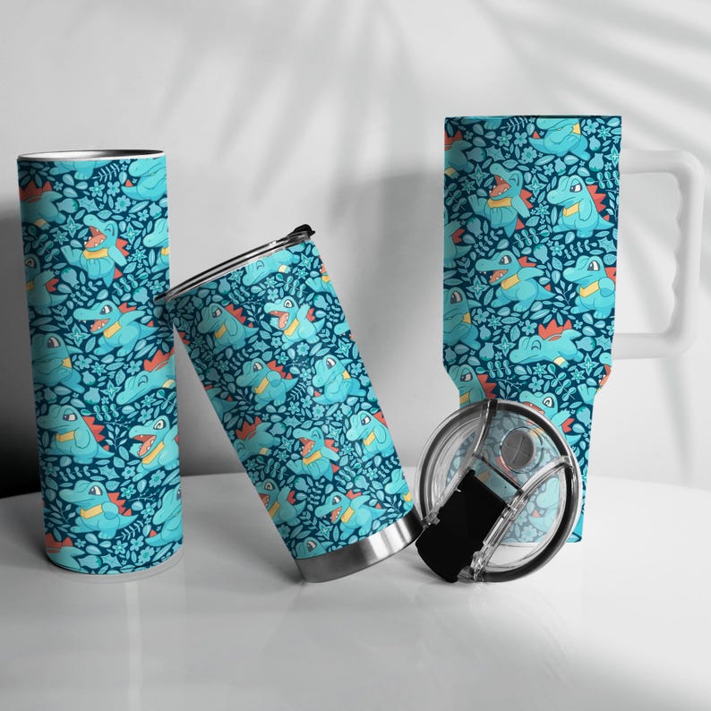 May include: A set of insulated travel mugs and a mug with a repeating pattern of cartoon alligators in blue, orange, and yellow on a dark teal background. The mugs are cylindrical, and the mug has a white handle.