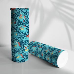 May include: Two cylindrical tumblers. The tumblers feature a teal background with a repeating pattern of cartoon alligators, flowers, and leaves. One tumbler stands upright, while the other lies on its side. The alligators are light blue with yellow and red accents.