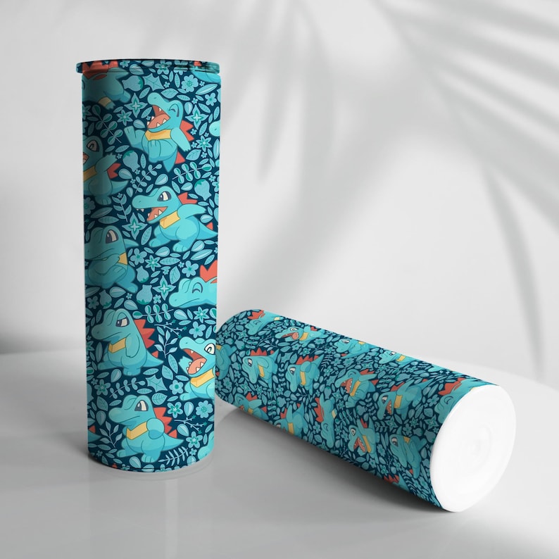 May include: Two cylindrical tumblers. The tumblers feature a teal background with a repeating pattern of cartoon alligators, flowers, and leaves. One tumbler stands upright, while the other lies on its side. The alligators are light blue with yellow and red accents.