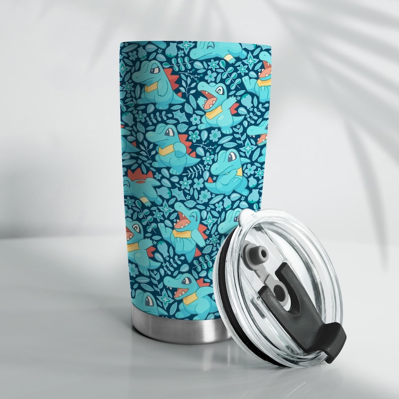May include: A stainless steel travel mug with a teal and blue pattern of cartoon alligators and floral designs. The lid is clear with a black handle. The mug is suitable for hot or cold drinks.