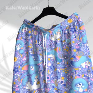 May include: Light purple shorts with a whimsical pattern featuring cartoon unicorns, mushrooms, stars, and musical notes. The shorts have a drawstring and are hanging on a black hanger. The design includes shades of blue, pink, yellow, and white.