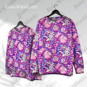 Könnte beinhalten: Zwei Sweatshirts mit Rundhalsausschnitt und einem lebendigen Muster aus rosa und blauen Zeichentrickfiguren und Blumen auf lila Grund. Die Sweatshirts hängen an schwarzen Kleiderbügeln.