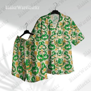 Puede incluir: Conjunto a juego de camisa de manga corta y pantalones cortos. El conjunto presenta un patrón repetido de dragones verdes y detalles florales amarillos sobre un fondo blanquecino. La camisa tiene cuello en V y los pantalones cortos tienen cintura con cordón.