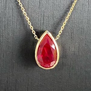 Puede incluir: Un collar de oro con un colgante de piedra preciosa roja en forma de lágrima. El colgante está engastado en un bisel de oro y cuelga de una delicada cadena de oro. La piedra preciosa tiene un color rojo vibrante.